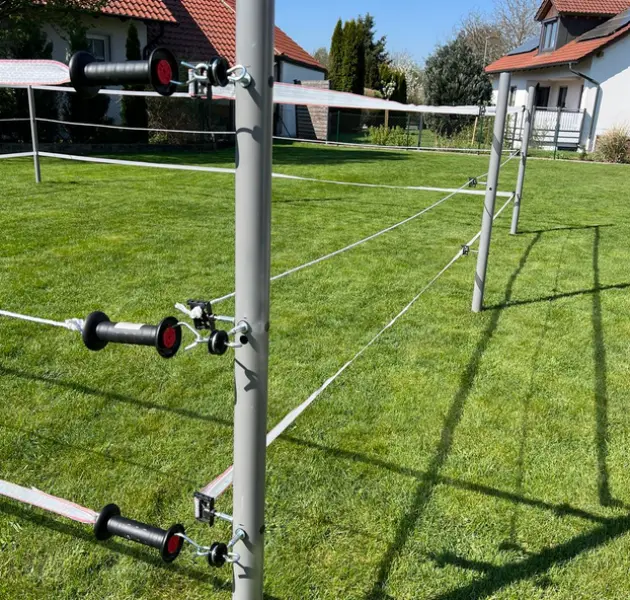 Elektrozaun-Drähte an Metallpfosten umschließen einen grasbewachsenen Hof, im Hintergrund sind Häuser zu sehen.